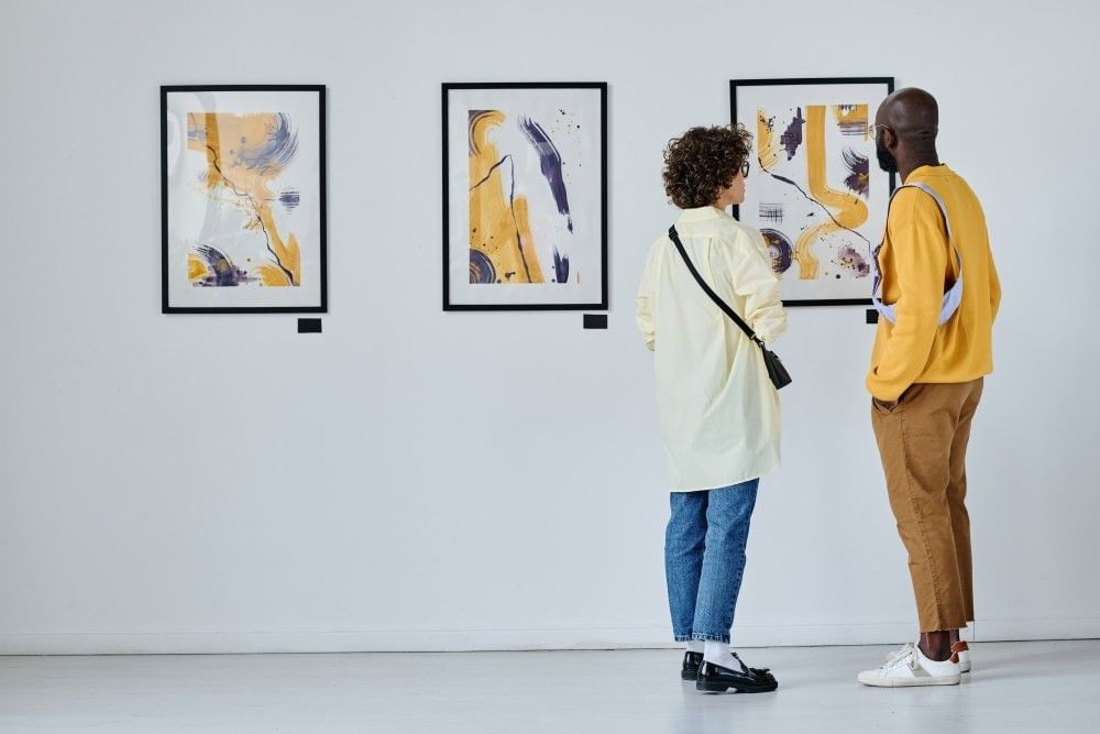 A young couple stand in an art gallery looking at three paintings that are black, white, and yellow.