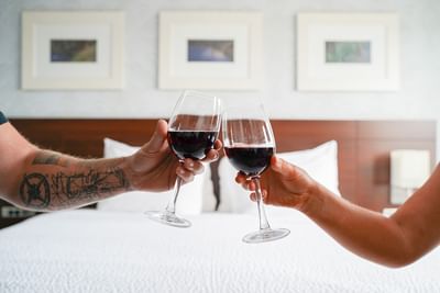 Close-up of two guests clinking glasses of red wine over a white king bed with large pillows at Rodd Moncton
