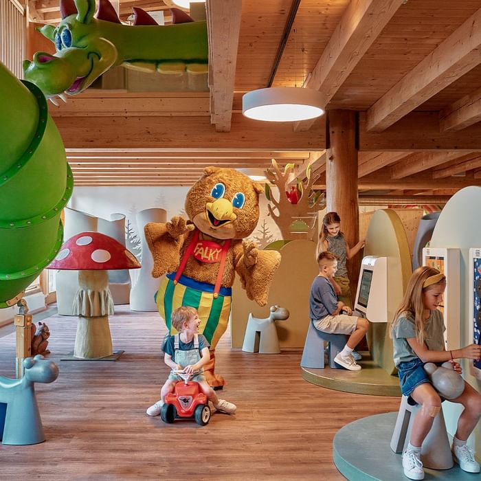 Kinder in einem Spielbereich mit Holzboden, Maskottchen und bunten Tierfiguren.