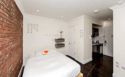 East Village Hotel room with brick wall, white bed, two floating shelves, and adjacent kitchenette with dark hardwood floors.