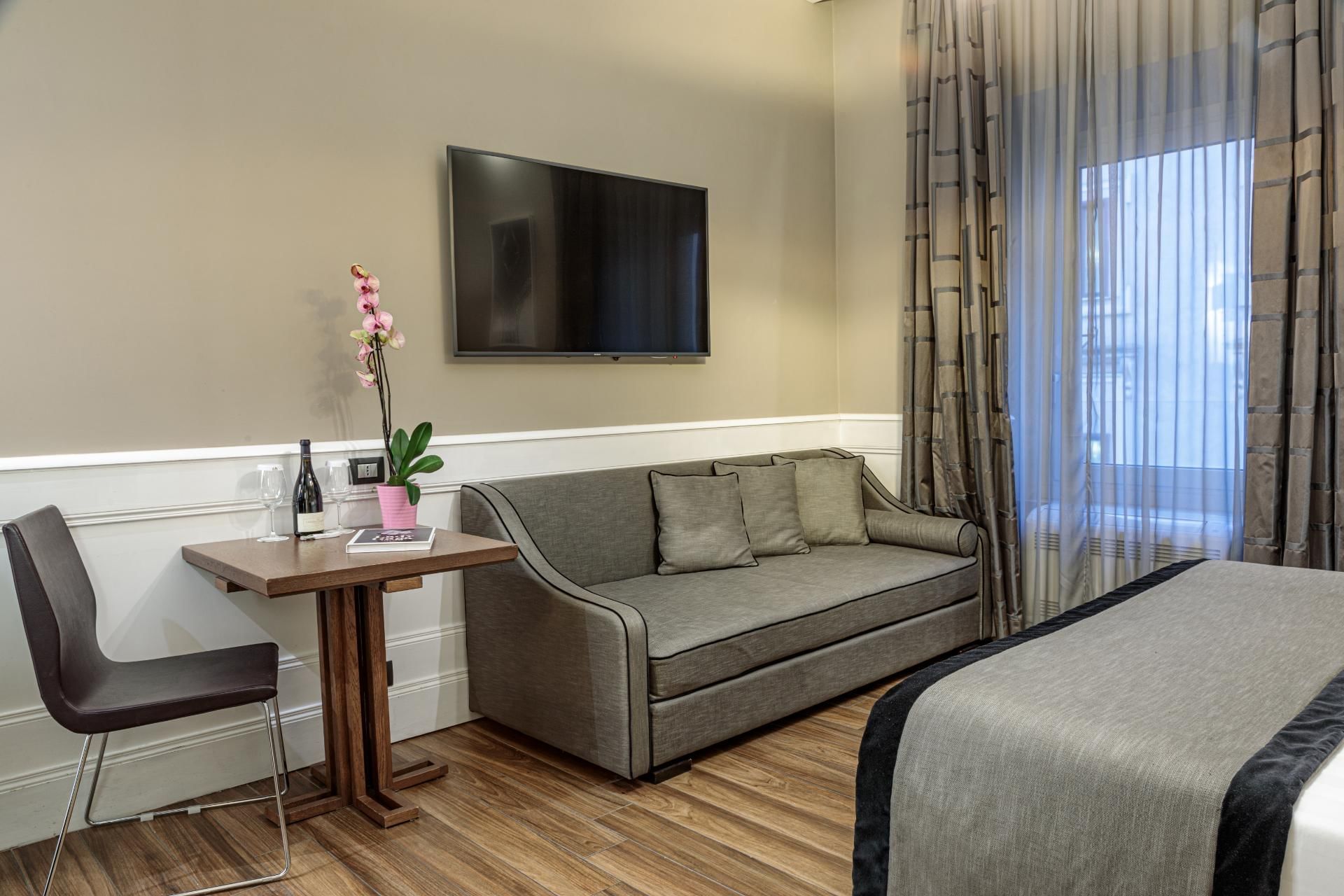 Stylish sofa, wooden table with a wine bottle, and a wall-mounted TV in the Deluxe Room at The Independent Suites