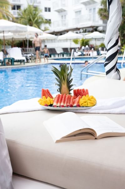 Tray of fruit and an open book on a poolside lounger with umbrellas and chairs at The Savoy Hotel & Beach Club