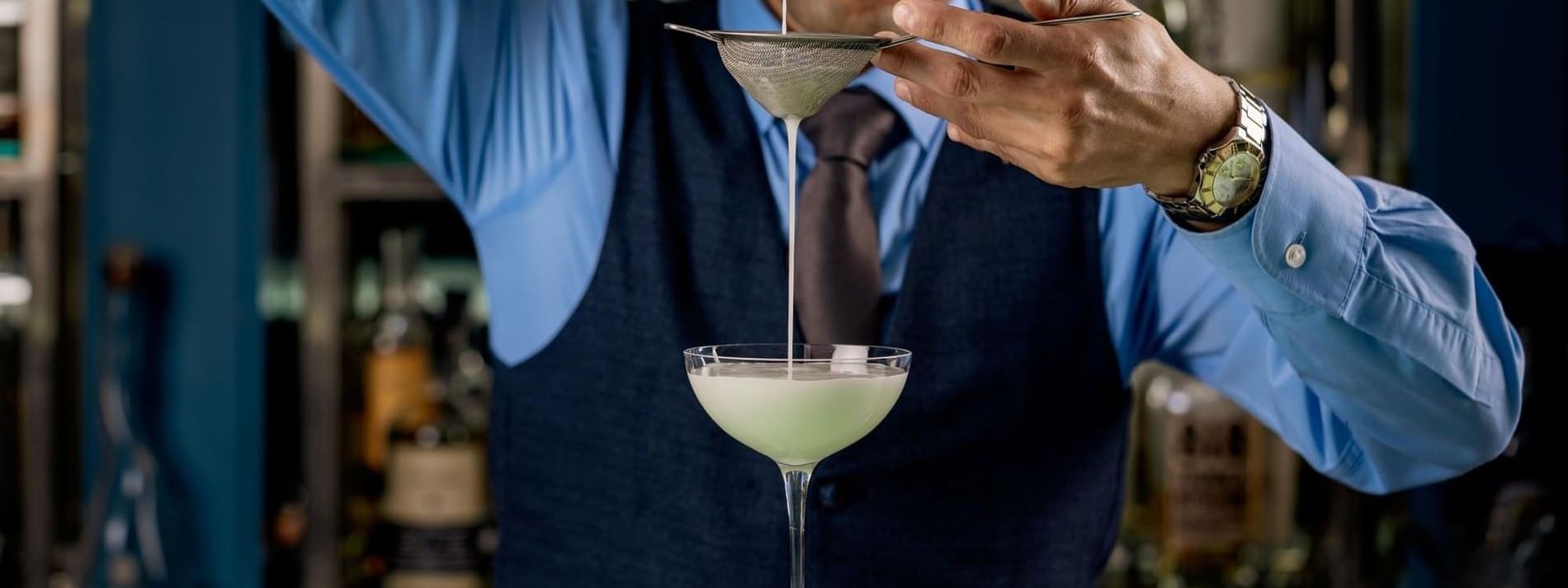 Bartender pouring a pale green cocktail through a strainer into a coupe glass in the bar in Tom Brown at The Capital