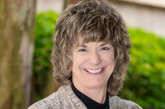 Kim DeBord with short layered brown hair in front of blurred green foliage backdrop at Rosen Inn at Pointe Orlando