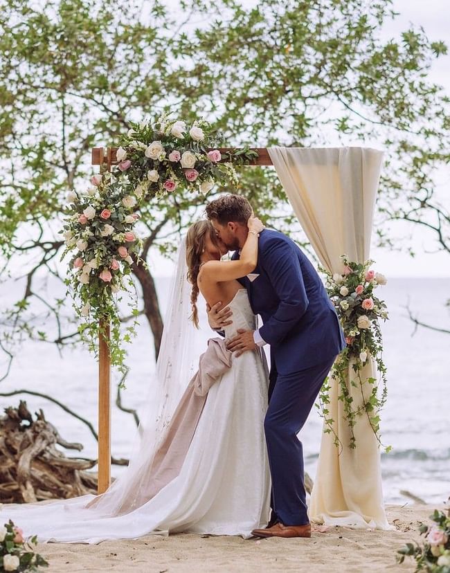 Beach wedding ceremony featuring a couple's first kiss under a floral wooden arch near Cala Luna Boutique Hotel