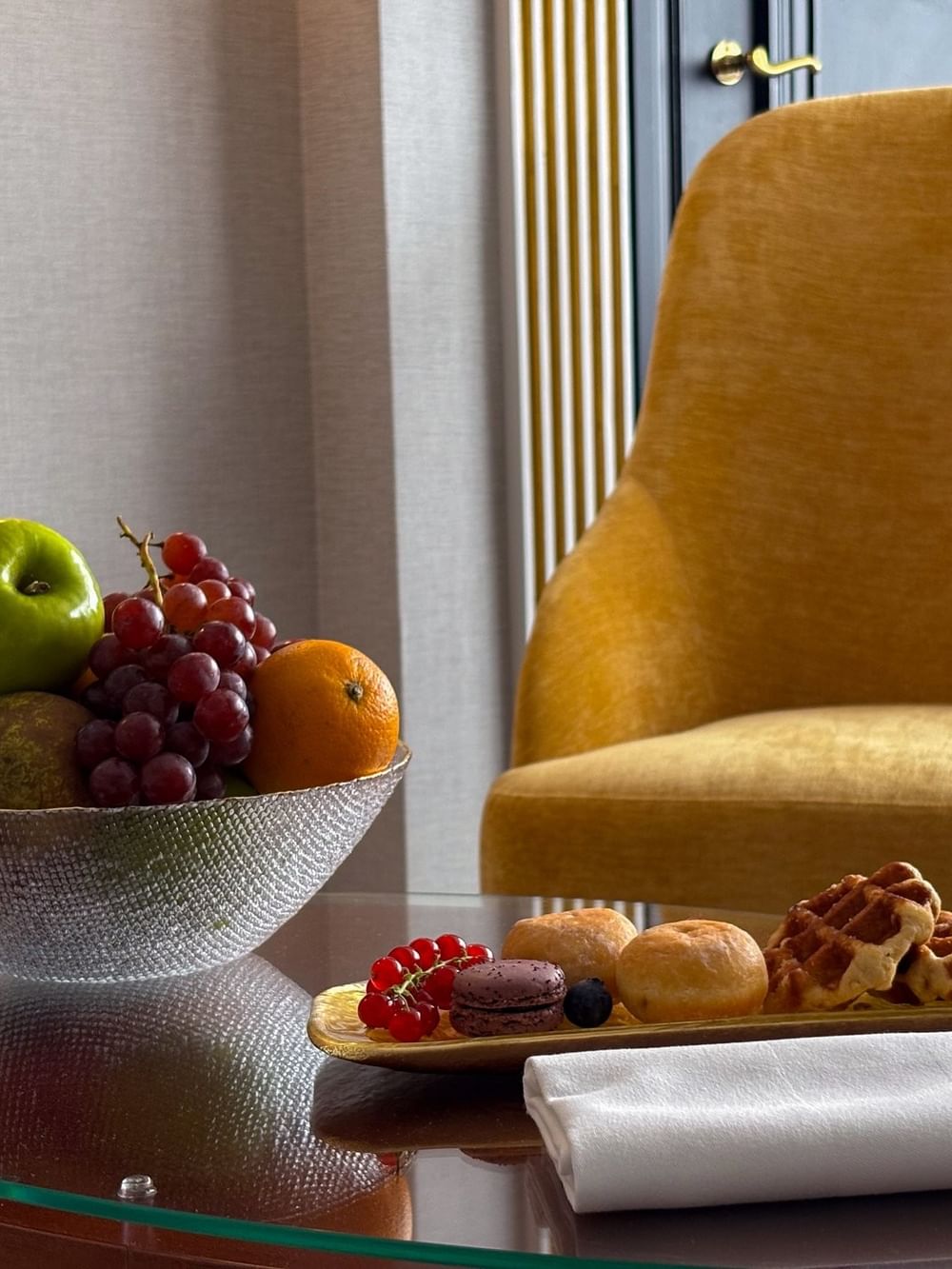 Een fruitschaal en gebakjes op een glazen tafel met een gele stoel in Hotel Barsey by Warwick.