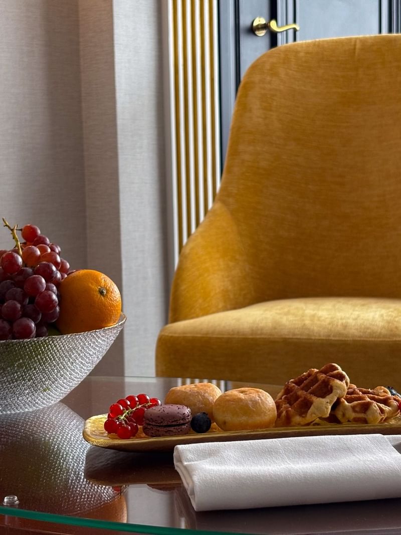 Een schaal met fruit en gebak op een glazen tafel met een gele stoel in Hotel Barsey by Warwick.