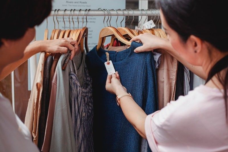 Two women browsing clothes in a store, with one woman examining the price tag on a blue dress near Real Inn Nuevo Laredo