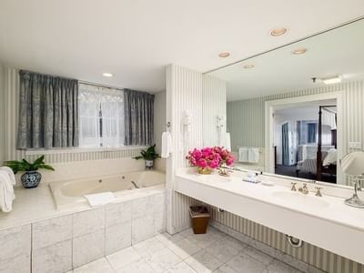Jetted tub by curtained window, under a pink flower arrangement near a double vanity with mirror at Simsbury 1820 House