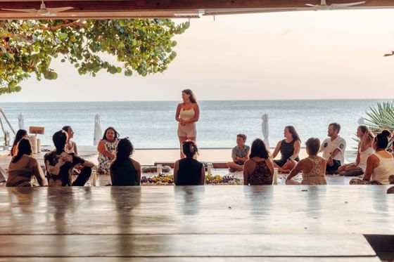 A large group of people sits in a circle on a wooden deck as a woman speaks, a wellness retreat at Ibagari Hotel