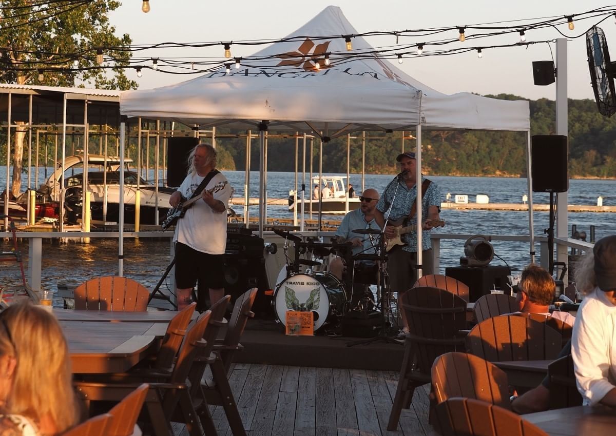 A live three-piece band performs under a white tent on a wooden deck by the water at Shangri-La Resort and Golf Club