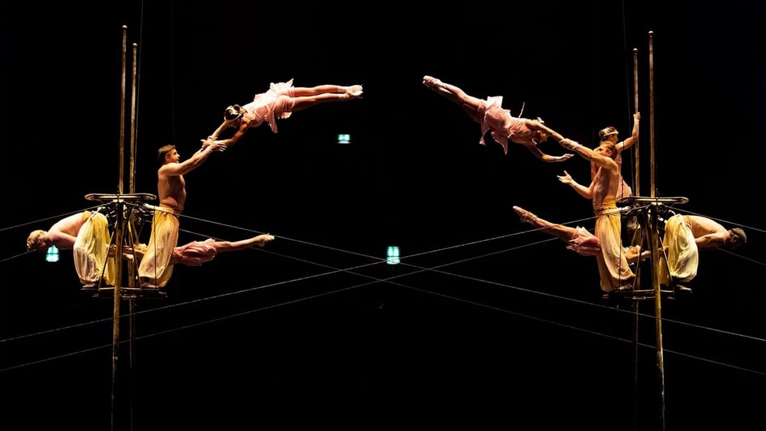 Acrobatic performers suspended in the air on trapezes, captured in a mirrored effect on a dark stage.