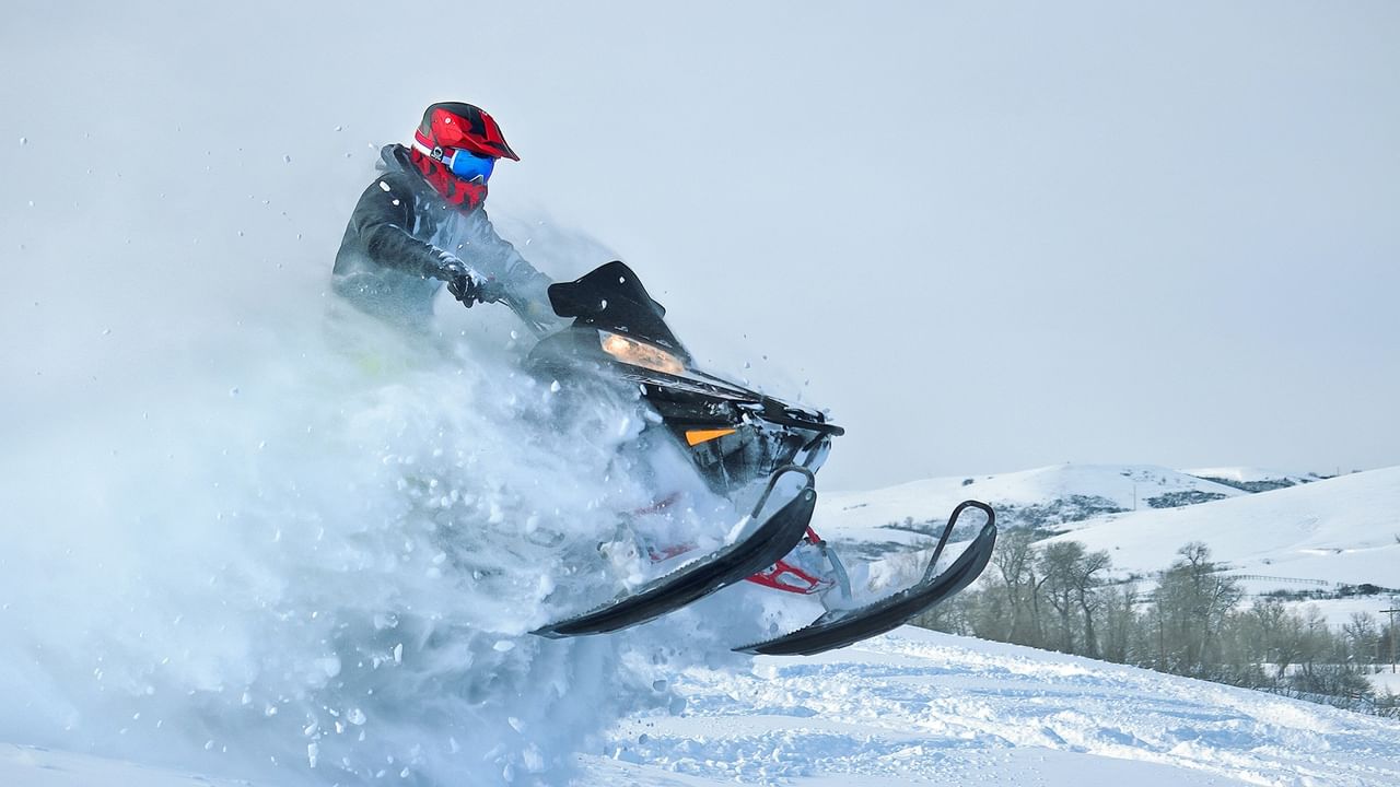 Person riding a snowmobile through snow with a red helmet and goggles, and snowy mountains in the background.