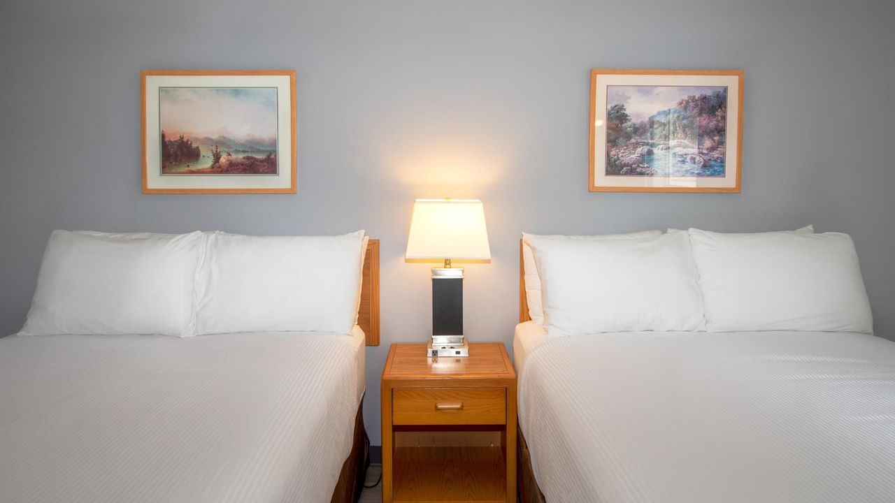 Two beds with white linens, a nightstand with a lamp, and two framed paintings on the wall.