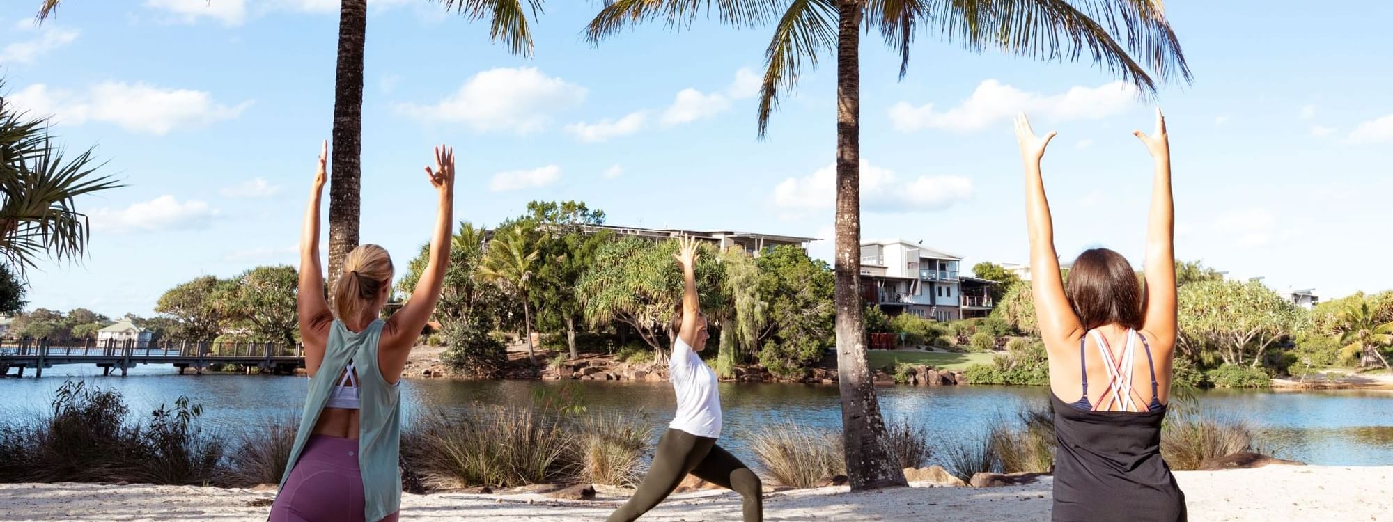 Three women practicing yoga on sand near a lake with palm trees under a bright sky at Novotel Sunshine Coast Resort