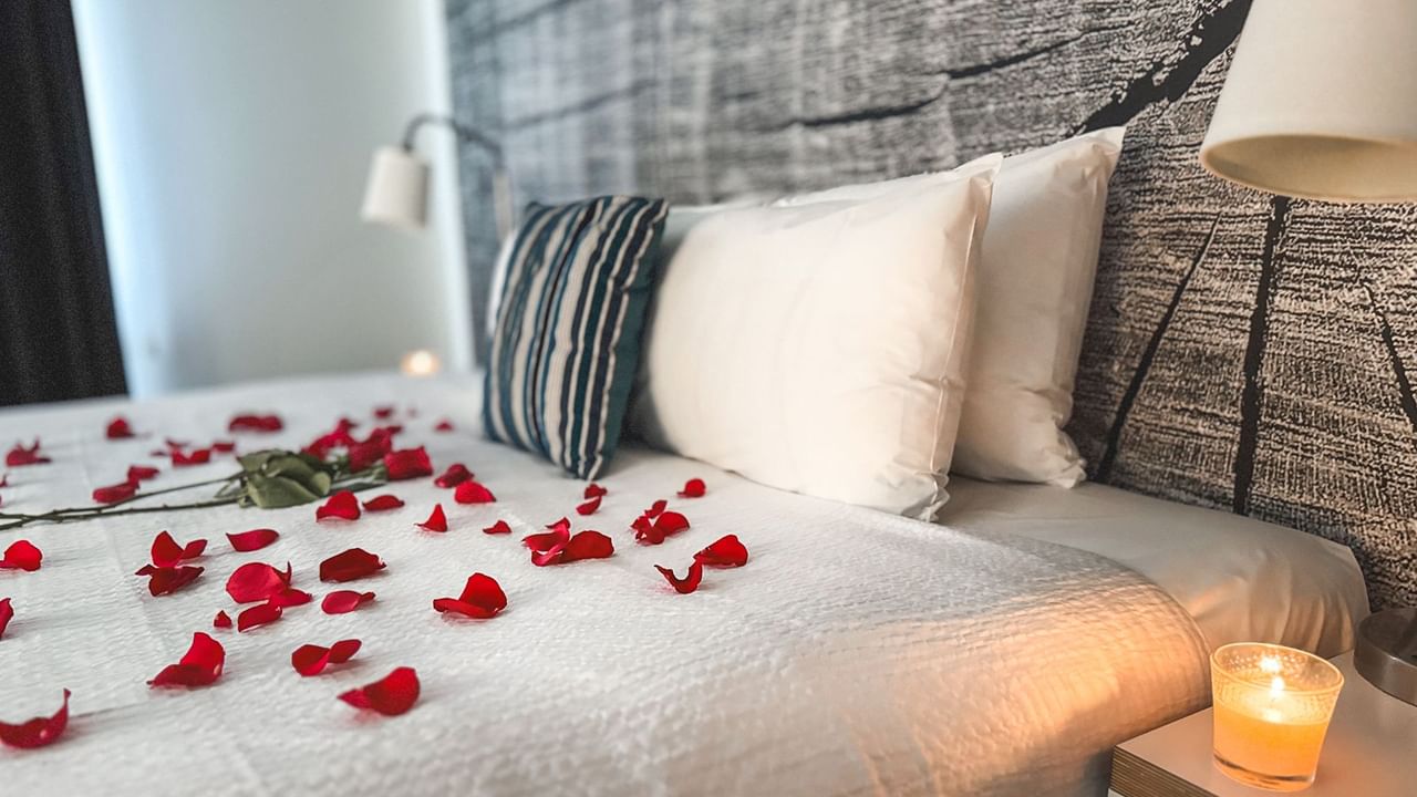 Bed with rose petals, and a lit candle on a nightstand with a lamp.