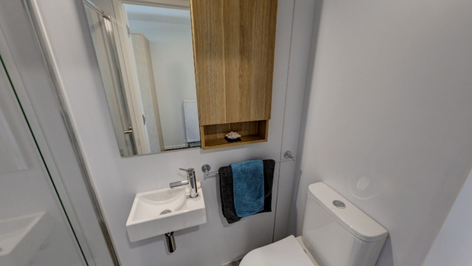 Compact modern bathroom with white sink, mirror, wooden cabinet, and blue and black towels in Tupuārangi