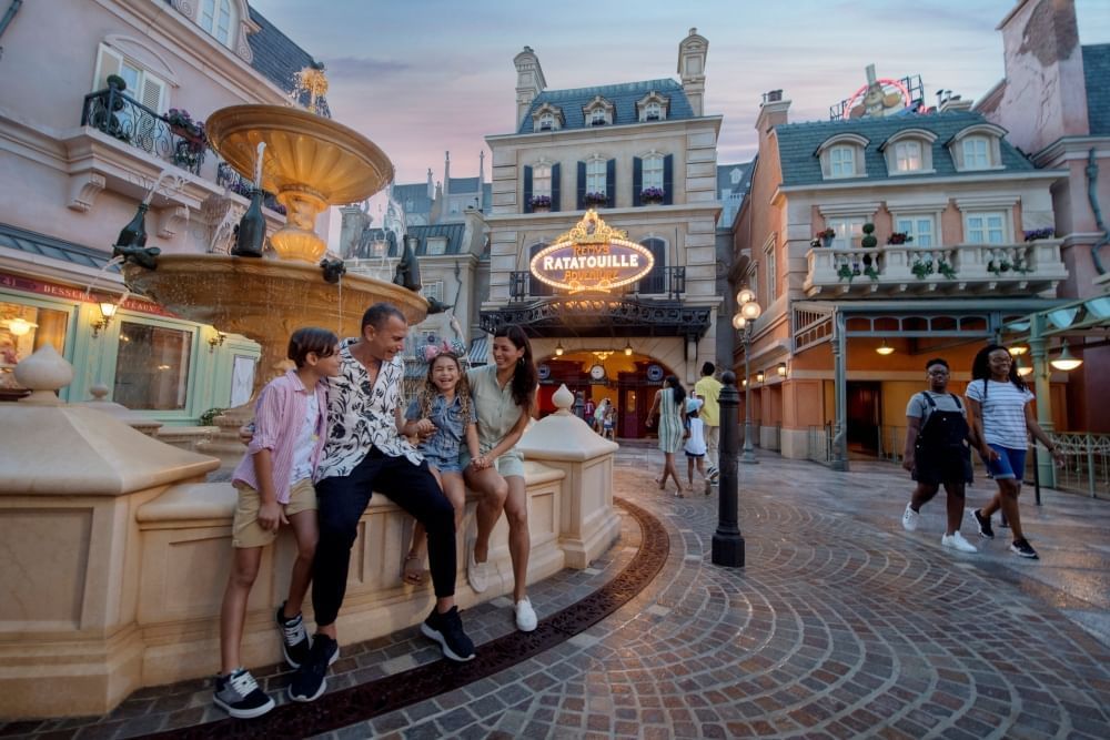 A family of four sits on the side of a fountain at twilight, surrounded by Parisian buildings, one with a lit sign that reads Ratatouille. 