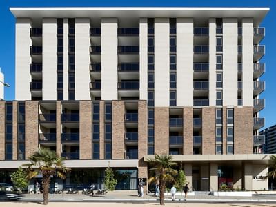 Modern multi-story building with large windows and balconies, set against a clear blue sky.