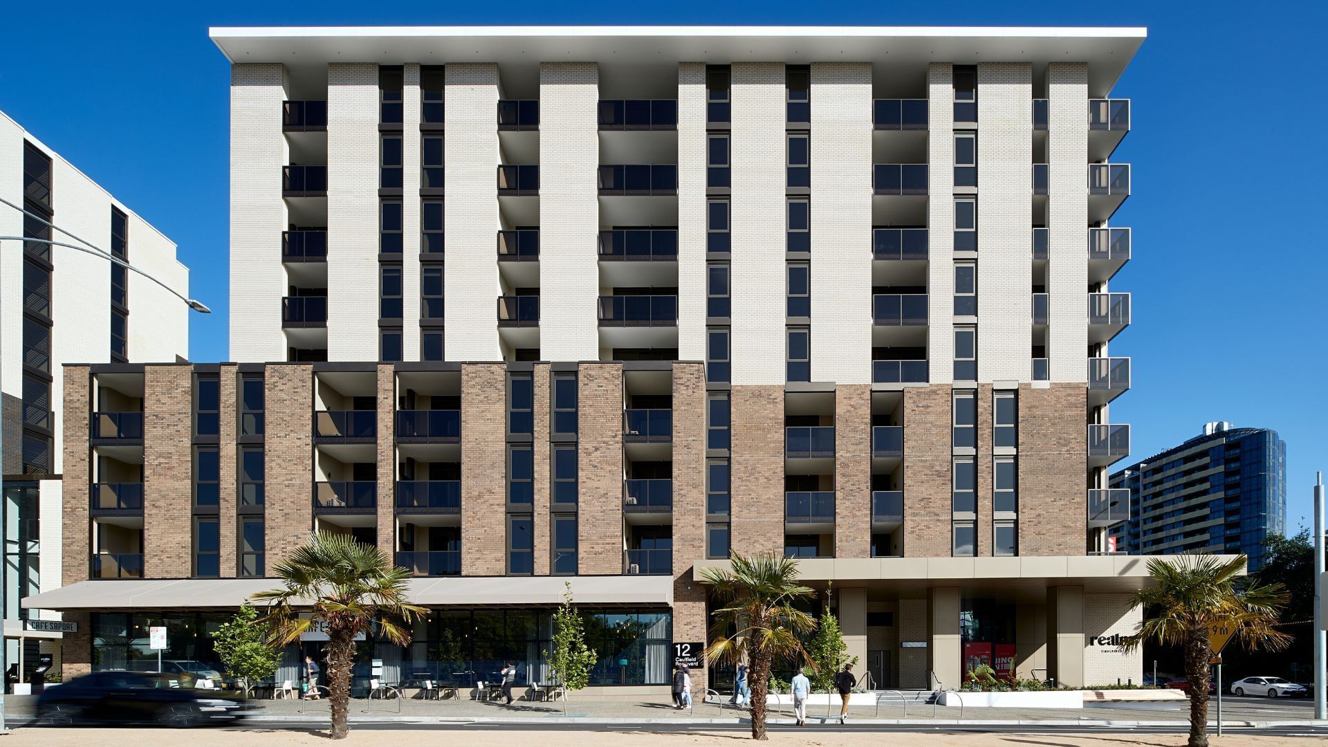 Modern multi-story building with large windows and balconies, set against a clear blue sky.