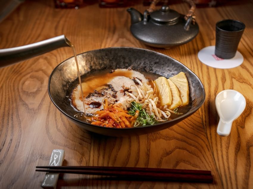 Cuenco de Ramen al estilo Morimoto con palillos y tetera sobre mesa de madera.