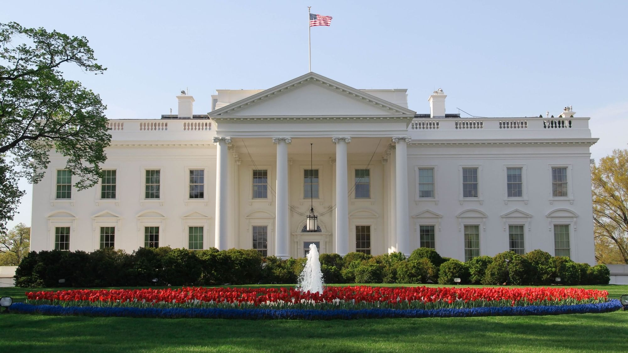 The front of a large white building with columns and a flag, surrounded by flowers and a fountain.