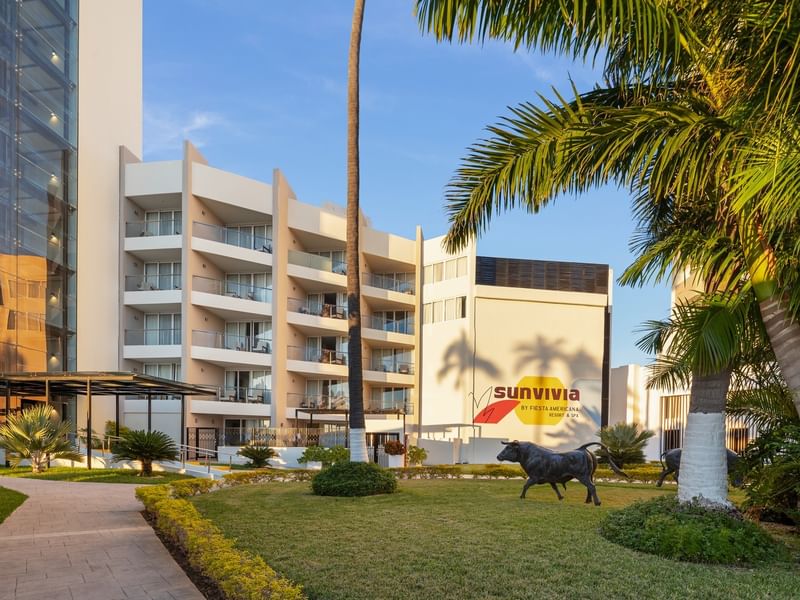 Exterior view of Sunvivia Mazatlán resort with a green lawn, palm trees, and a bronze bull statue near the entrance