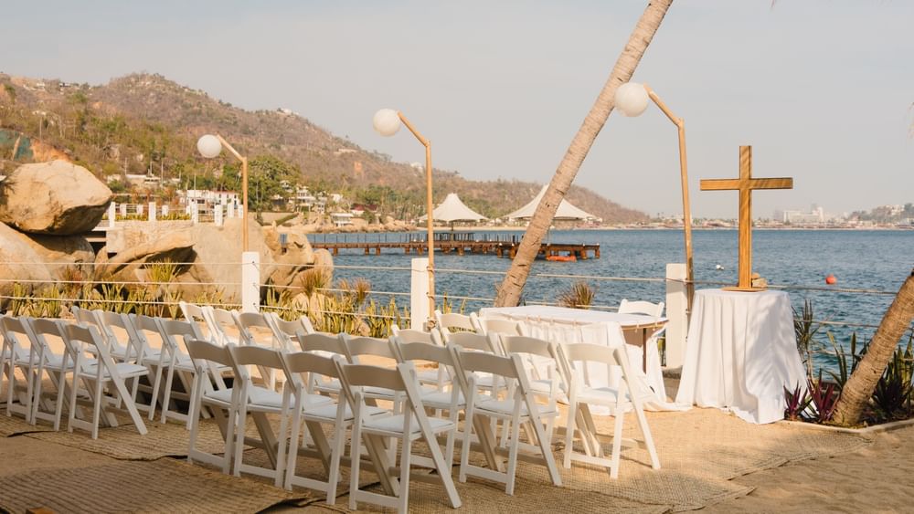 Outdoor wedding ceremony setup with white chairs and a wooden cross by the sea at Camino Real Acapulco Diamante