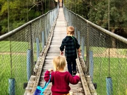 Boy and girl walk together across a wooden suspension bridge over a green field near The Sebel Mandurah