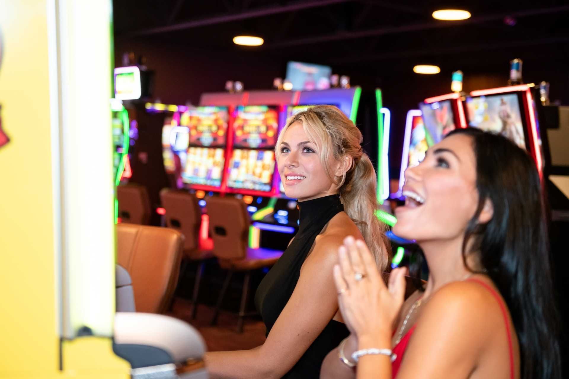Two guests enjoy casino gaming machines by rows of colorful illuminated slots under ceiling lights at The Brook