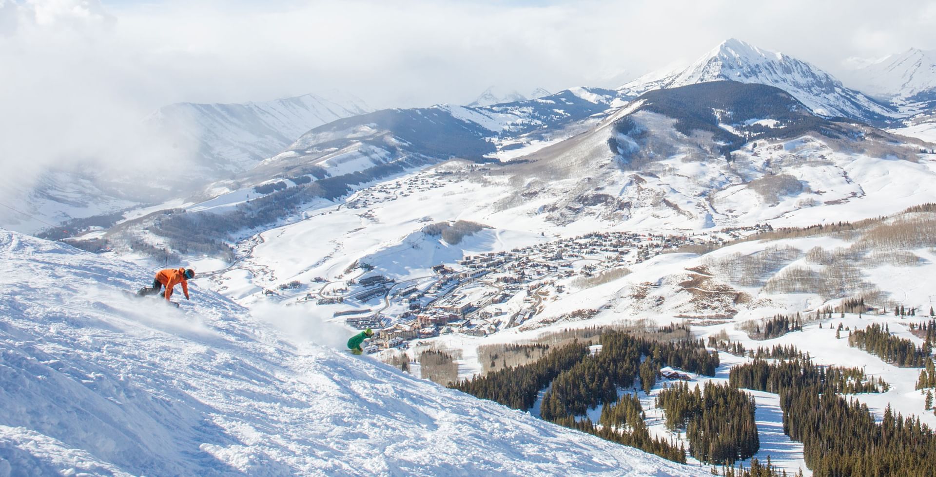 Two snowboarders navigating snowy mountain slopes at Elevation Hotel in Crested Butte Colorado  with a town and lush forest.