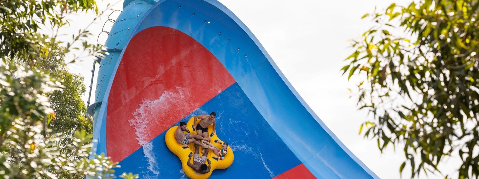 Four people on a yellow raft slide down a blue and red water slide surrounded by trees.