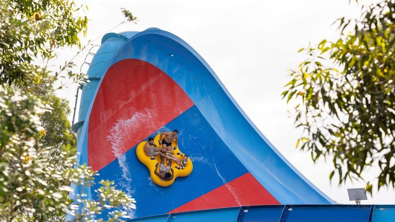 People on a yellow tube sliding down a blue and red water slide with trees in the background.