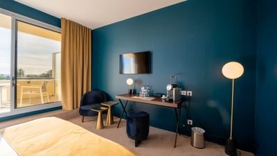 A stylish hotel room with a bed, blue chair, desk, TV, and a balcony with outdoor furniture.