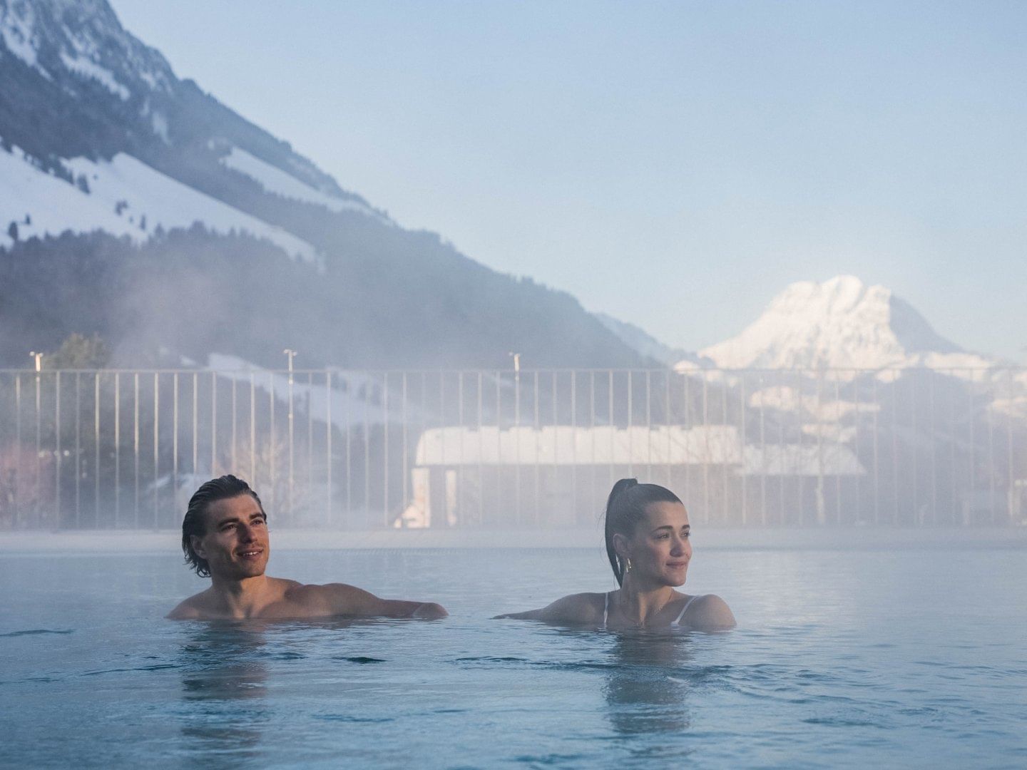 Couple relaxing in Les Bains De La Gruyere pool surrounded by snow-capped mountains near Hotel des Innovations