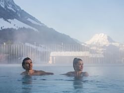 Couple relaxing in Les Bains De La Gruyere pool surrounded by snow-capped mountains near Hotel des Innovations