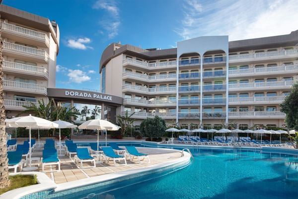A large resort hotel with white balconies overlooks a bright blue swimming pool lined with lounge chairs and umbrellas.