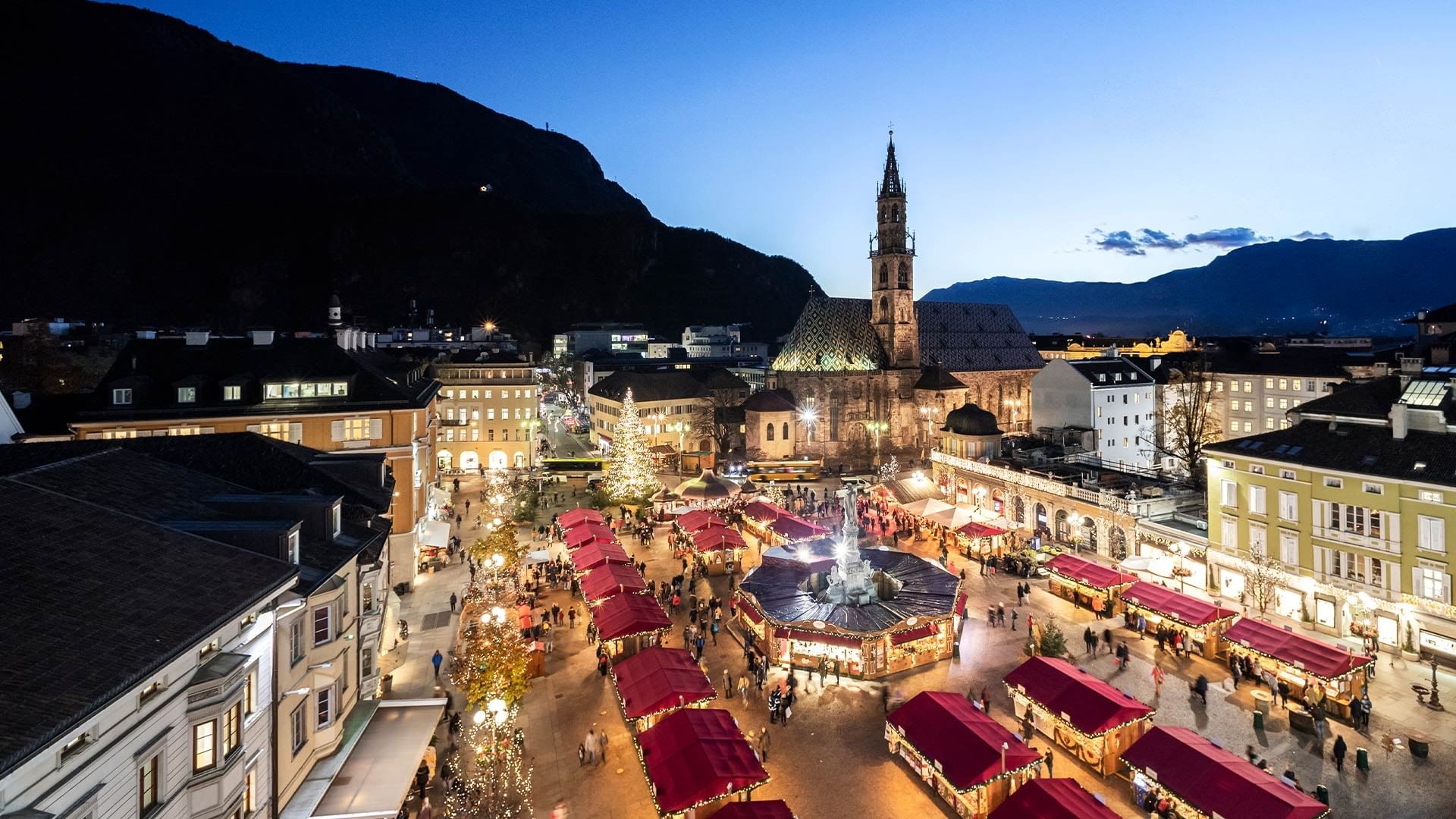 Luftaufnahme eines beleuchteten Marktplatzes mit Buden, umgeben von Gebäuden und einer Kirche.