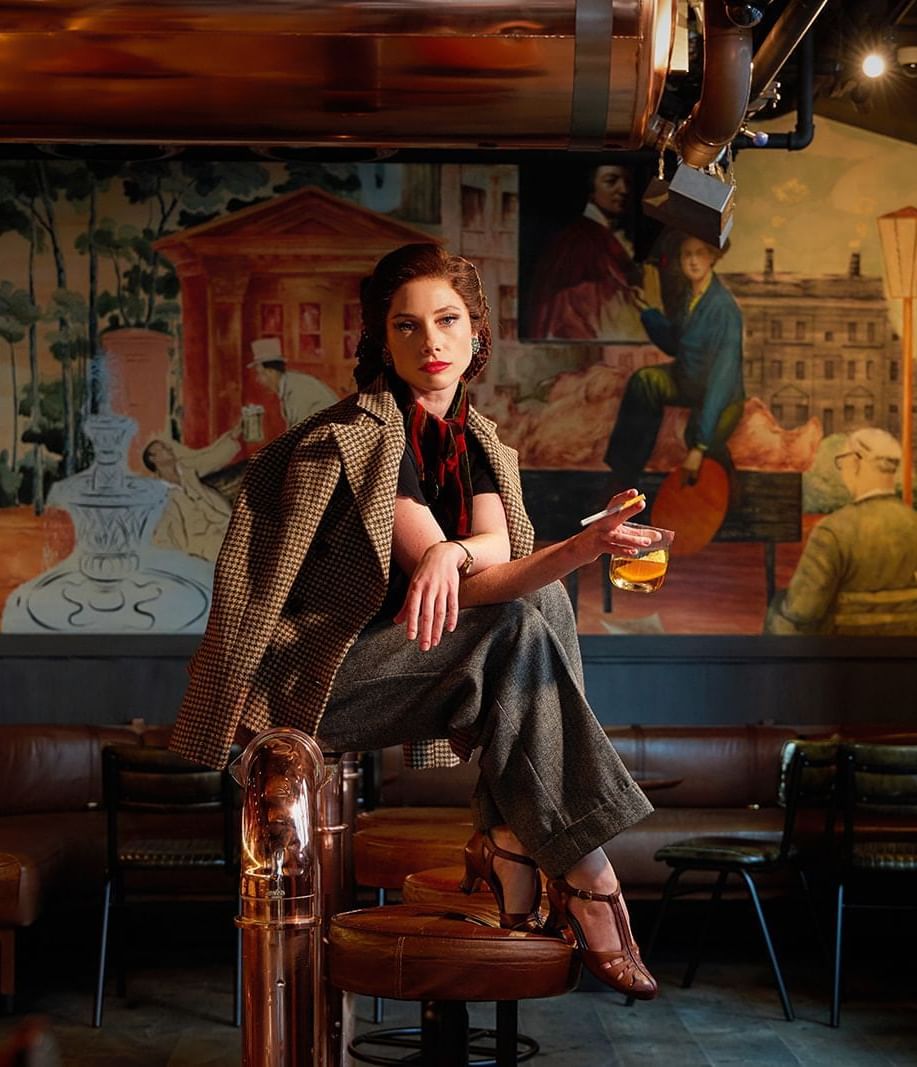 A stylish woman posing with a drink in a sophisticated Midnight Bell bar with a mural at The Londoner Hotel