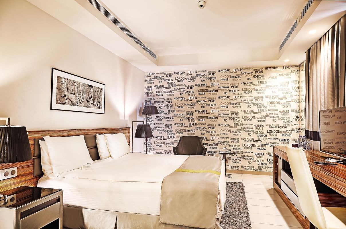 Bed by a desk under city-themed wallpaper near a grey armchair in Deluxe Room at Warwick Stone 55 - Beirut