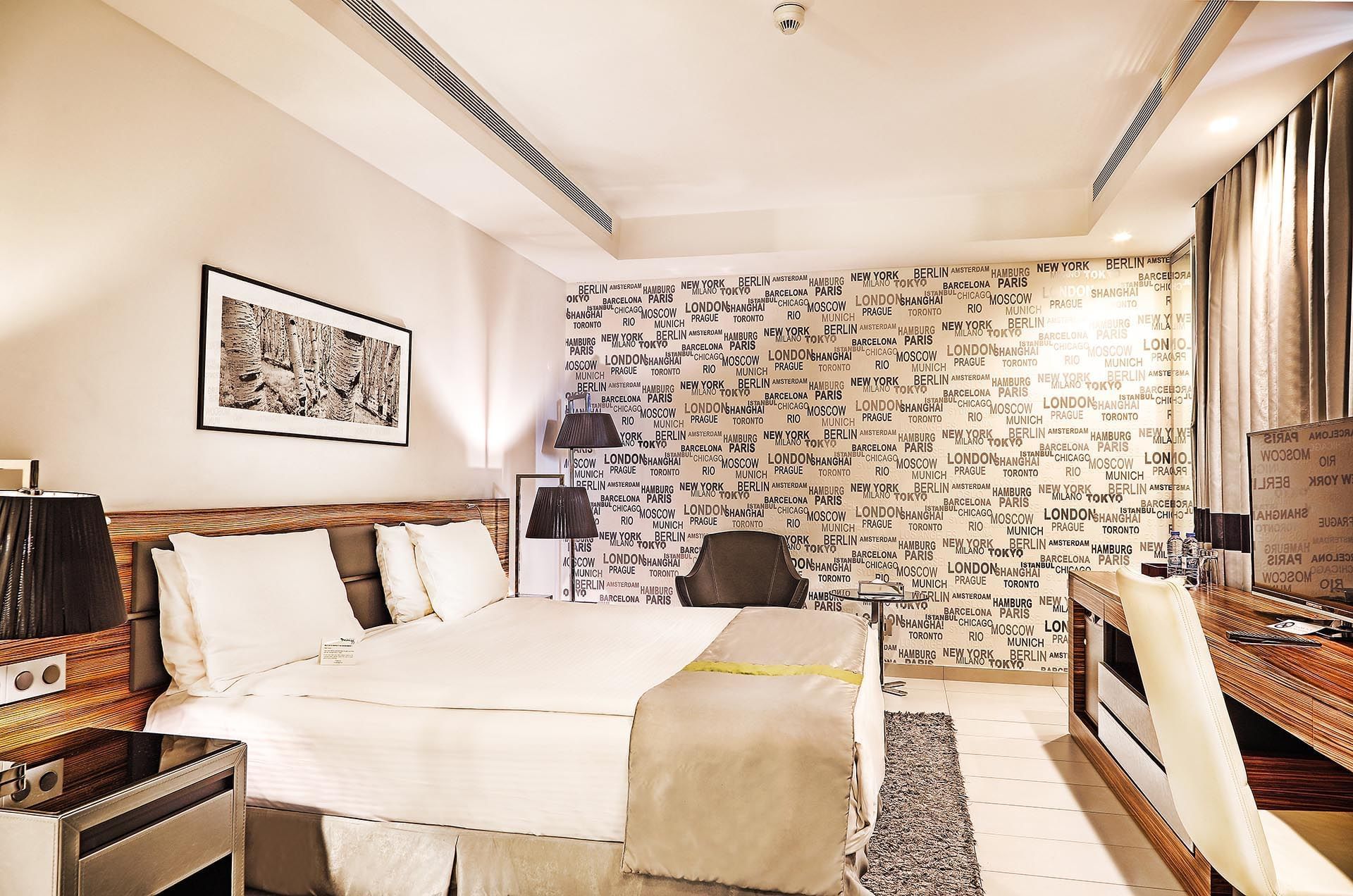 Bed by a desk under city-themed wallpaper near a grey armchair in Deluxe Room at Warwick Stone 55 - Beirut