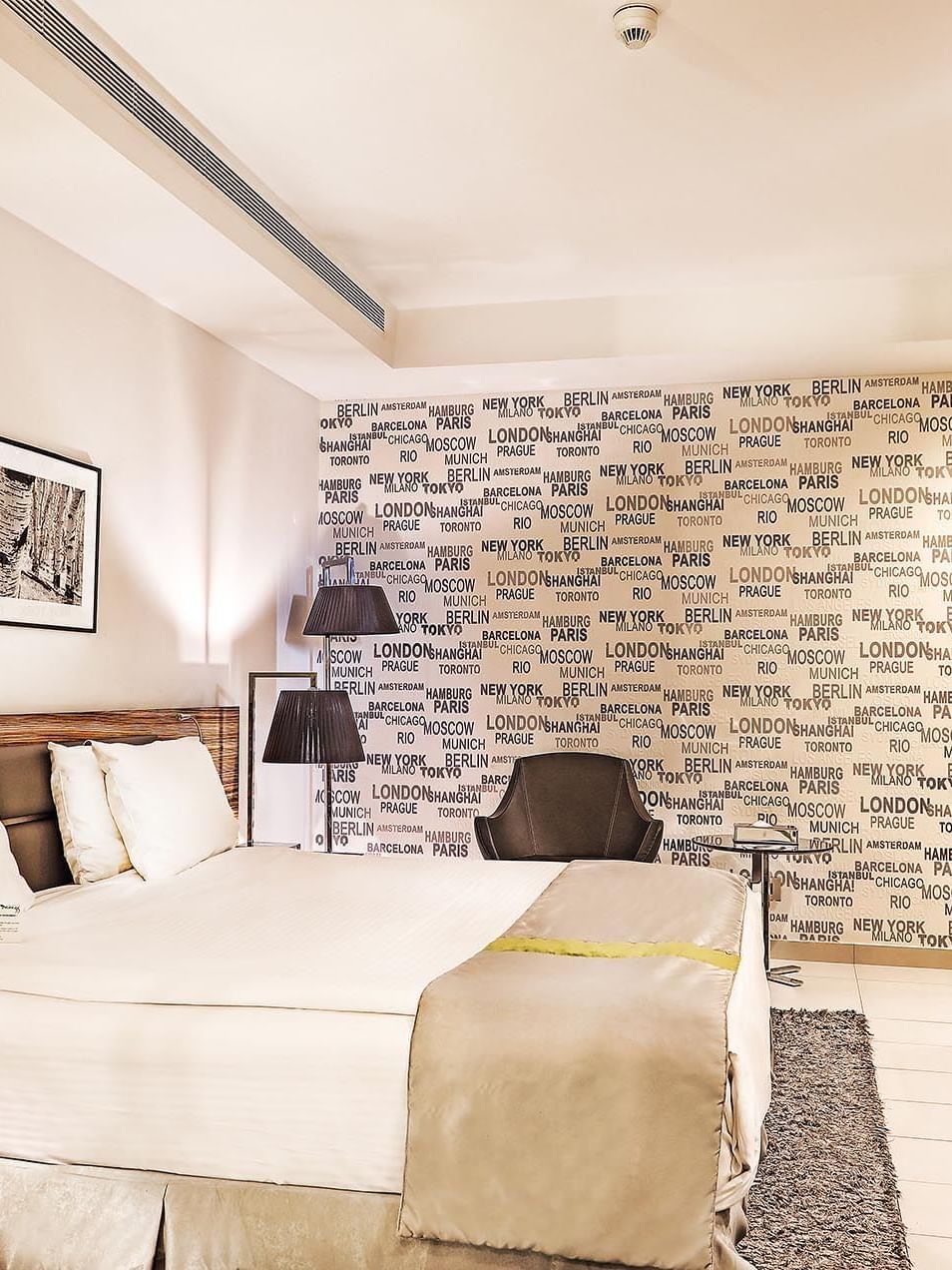 Bed by a desk under city-themed wallpaper near a grey armchair in Deluxe Room at Warwick Stone 55 - Beirut