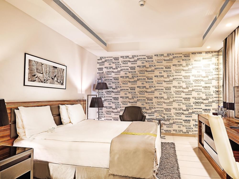 Bed by a desk under city-themed wallpaper near a grey armchair in Deluxe Room at Warwick Stone 55 - Beirut