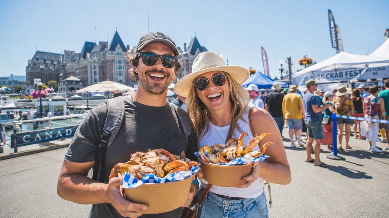 Smiling couple holding baskets of fresh crab at an outdoor waterfront festival with crowds and tents in the background.