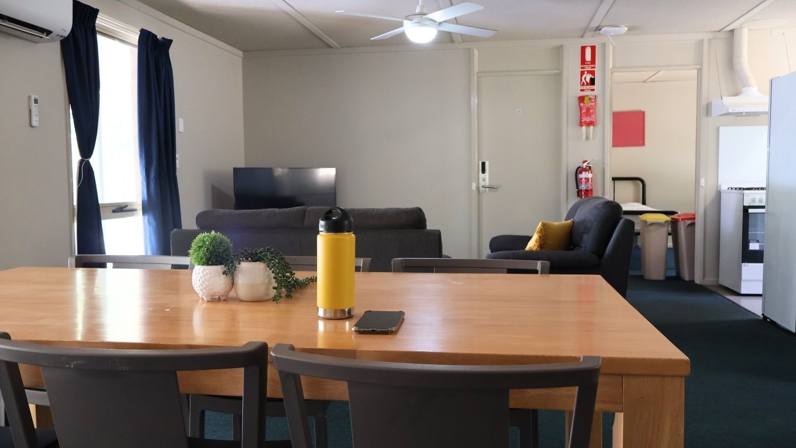Wooden dining table with plants, a water bottle, and a smartphone on top in a living room at La Trobe University Villas.