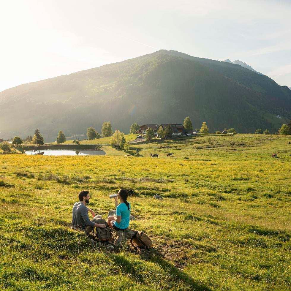Two people relaxing on a grassy field near a mountain with a pond in the background.