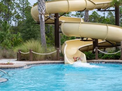 Child sliding down yellow water slide into outdoor pool surrounded by palm trees at Camp Creek Inn in Inlet Beach
