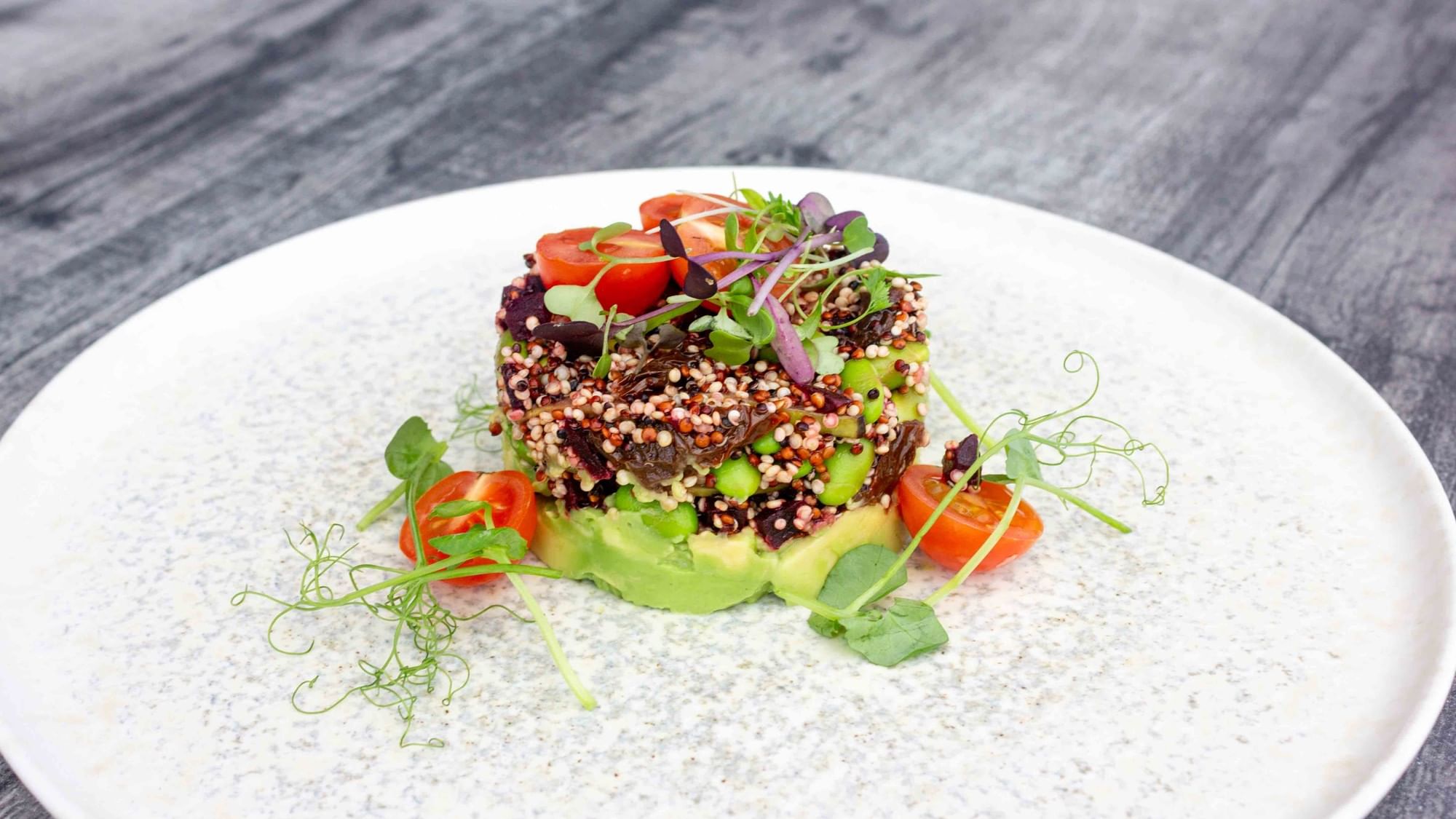 Plate with quinoa, avocado, green beans, and tomatoes garnished with herbs, served on a wooden table.
