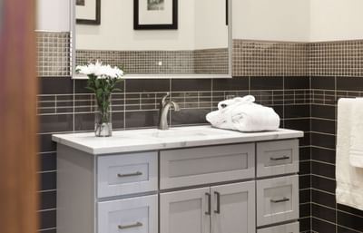 A modern bathroom with brown tiles, floral displays, hotel robe, light cabinetry, and a light-up mirror.