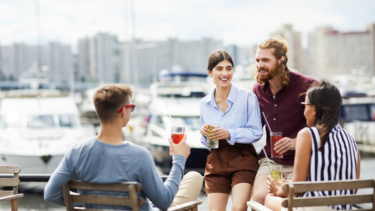 Friends enjoying drinks and conversation at a waterfront marina with boats and city buildings in the background.